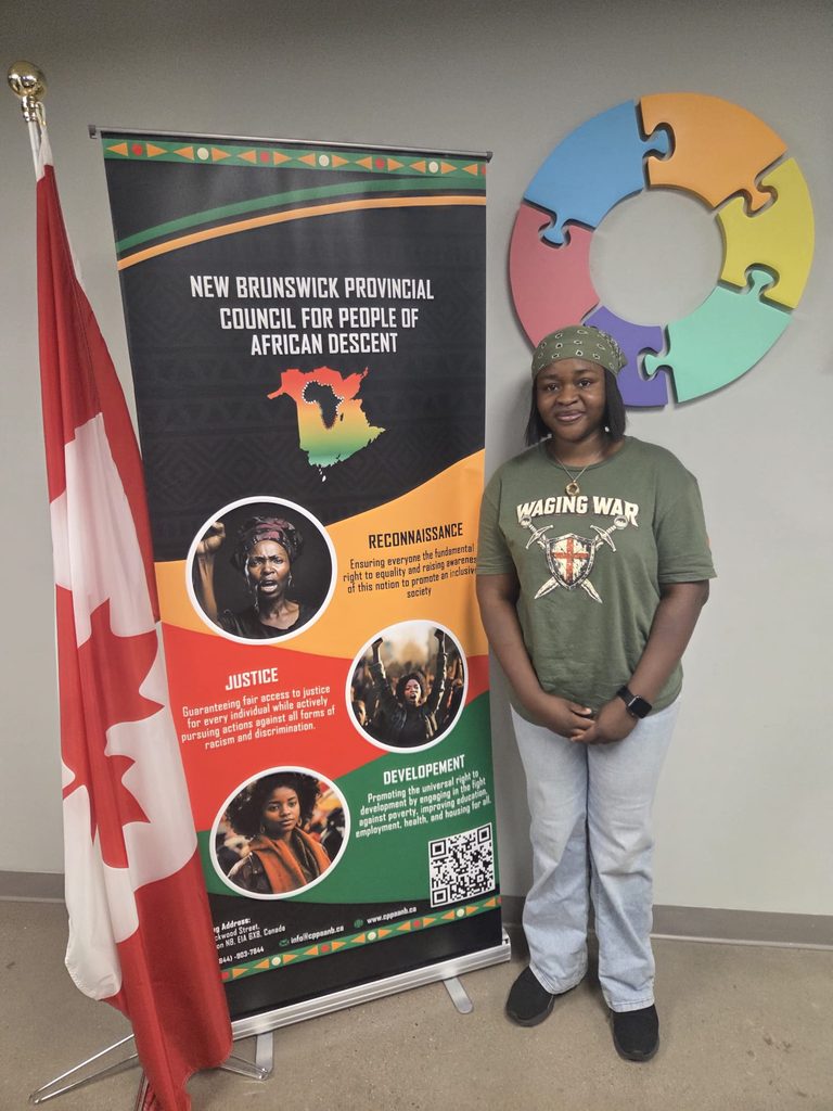 Oreoluwa Orokotan posing next to a New Brunswick Provincial Council for People of African Descent Event Banner